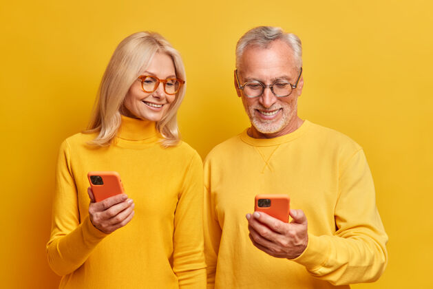 老年人上了年纪的奶奶和爷爷一起在智能手机设备上看照片 在网上看有趣有趣的视频 穿着休闲的黄色高领衫在室内摆姿势玩短信温柔