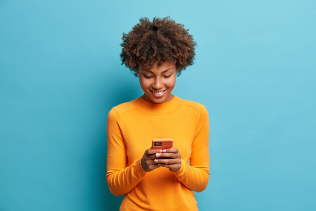 人快乐开朗的女人看着智能手机的屏幕享受在线聊天类型短信冲浪社交网络穿着随意的姿势对着蓝色的墙壁人跳投设备
