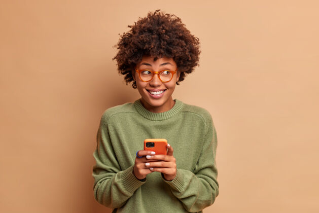沟通漂亮女人的画像用手机收到愉快的信息笑容满面 一边若有所思地看一边在社交网络隔离的米色墙上阅读她的帖子下的反馈眼镜民族快乐
