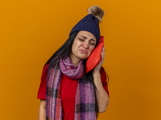 疾病悲伤的年轻白种人生病的女孩戴着冬天的帽子和围巾用热水袋抚摸着脸闭着眼睛隔离在橙色的墙上有复制空间触摸穿封闭