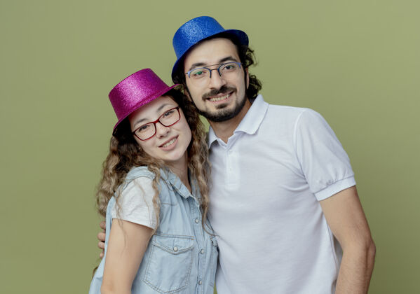 帽子微笑的年轻夫妇戴着粉红色和蓝色的帽子隔离在橄榄绿的墙上年轻情侣穿上