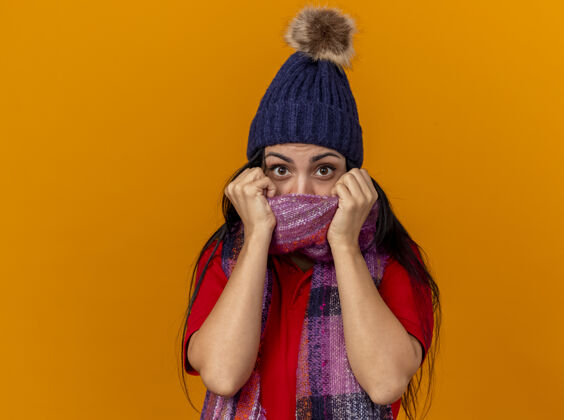 帽子印象深刻的年轻白种人生病的女孩戴着冬天的帽子和围巾 用围巾遮住嘴巴 隔离在橙色的墙上与复制空间橙色围巾穿着