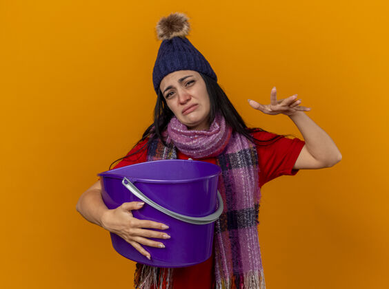帽子未被释放的年轻白种人生病的女孩戴着冬天的帽子和围巾拿着塑料桶有恶心保持手在空气中隔离在橙色墙上与复制空间围巾年轻穿
