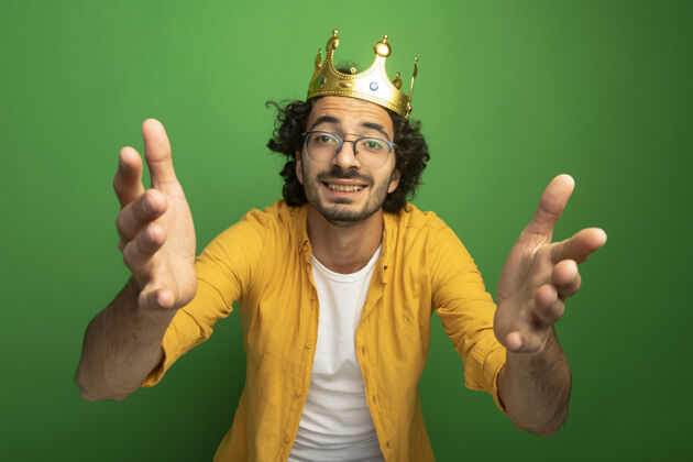 穿面带微笑的年轻英俊的白人男子戴着眼镜和皇冠伸出双手孤立在绿色的墙上伸展皇冠朝
