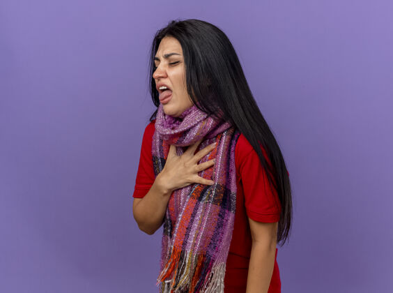 疾病恼怒的年轻白种人生病的女孩戴着围巾站在侧视图把手放在胸前展示舌头闭着眼睛隔离在紫色的墙壁上复制空间穿胸部关闭