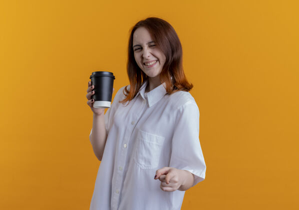 秀微笑着的年轻红发女孩拿着一杯咖啡 在黄色的墙上向你展示孤立的姿态微笑女孩杯子