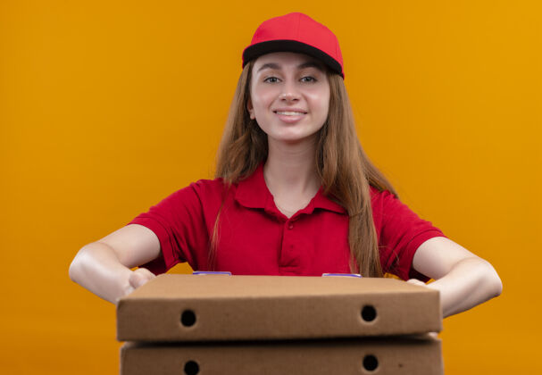 微笑微笑着的年轻送货女孩穿着红色制服 在孤立的橙色墙上伸展包裹盒子伸展分娩