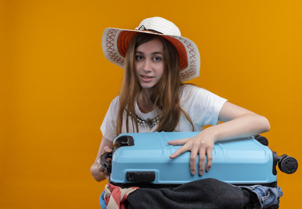 女孩年轻的旅行女孩戴着帽子 提着装满衣服的手提箱 看着孤立的橙色墙壁 还有复制空间帽子手提箱衣服