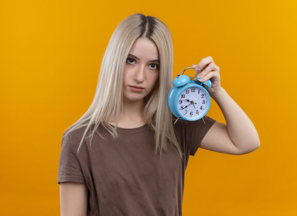年轻年轻的金发女孩在孤立的橙色墙上拿着闹钟金发女孩闹钟