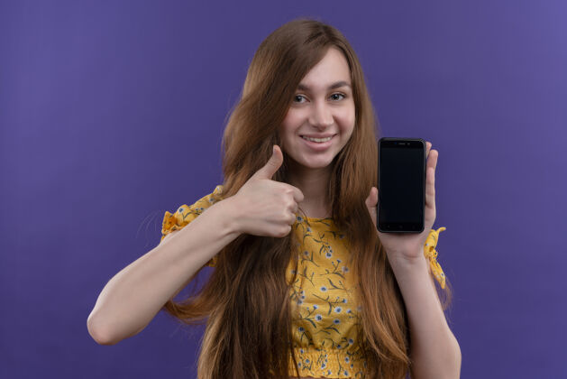 握着微笑的年轻女孩拿着手机 在孤立的紫色墙上竖起大拇指微笑表演电话