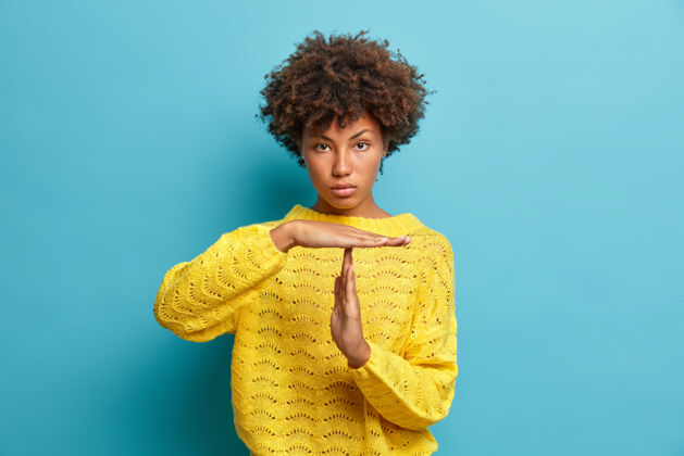 发型严肃自信的卷发女人做超时手势示范极限要求停止穿黄色针织套头衫隔离在蓝色墙上脸头发时尚