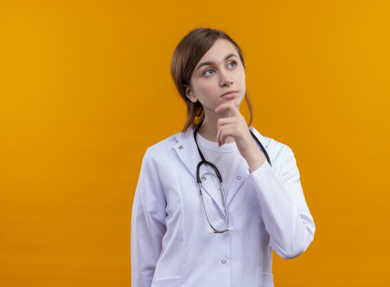 穿体贴的年轻女医生 穿着医用长袍 手放在下巴上的听诊器 放在隔离的橙色墙壁上 留有复印空间女人听诊器手