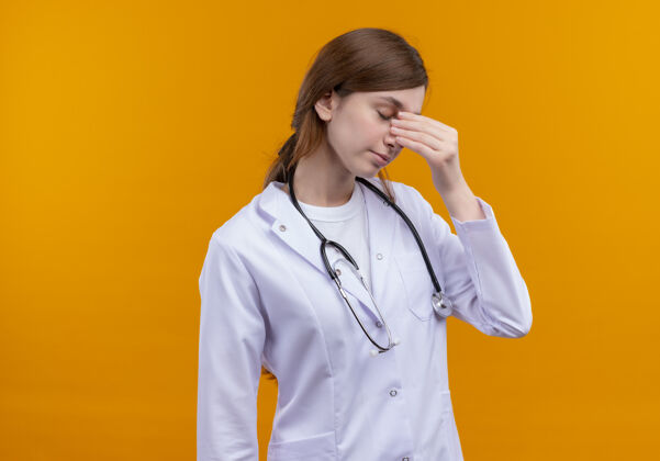 鼻子疲惫的年轻女医生穿着医用长袍和听诊器把手放在鼻子上孤立的橙色墙壁与复制空间听诊器长袍穿