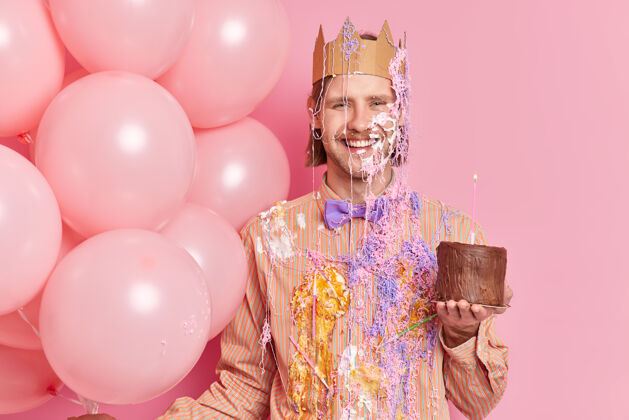 生日喜气洋洋的生日男士抹着奶油拿着巧克力蛋糕祝贺结婚纪念日有喜庆的心情在办公室里享受公司聚会的自由时光隔着粉色的墙壁束气球胡子