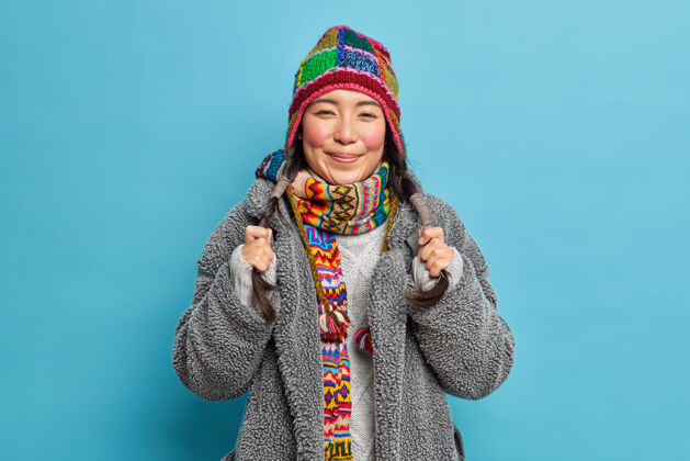 高兴亚洲女人的肖像拿着两个辫子微笑着愉快地穿着外套编织帽子和围巾围着脖子感觉快乐隔离在蓝色的墙壁上请成人外套
