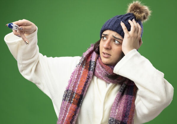 手关心的年轻白种人生病的女孩穿着长袍 戴着冬天的帽子和围巾 手放在头上 手举在绿色的墙上 看着温度计体温计穿疾病
