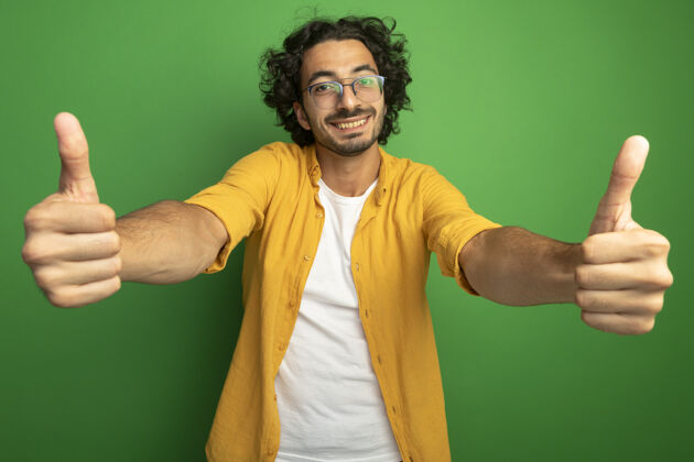 男人微笑着的年轻英俊的白人男子戴着眼镜 竖起大拇指孤立地站在绿色的墙上拇指帅哥年轻