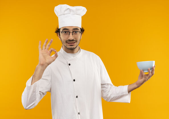 碗高兴的年轻男厨师穿着厨师制服 戴着眼镜 拿着碗 在黄色的墙上显示出奥基孤立的姿态手势好的制服