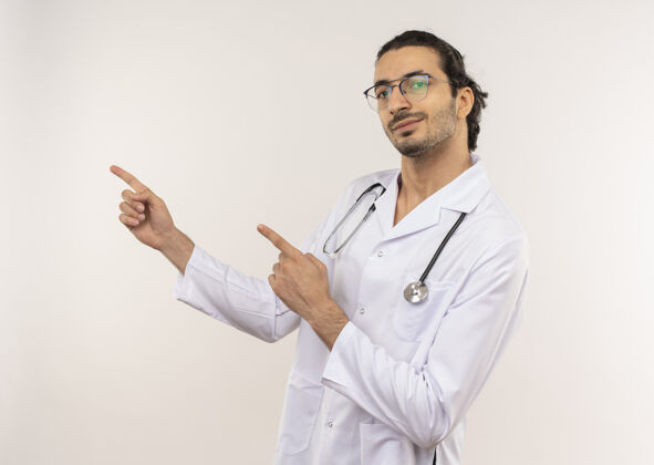 请戴高兴的年轻男医生戴着眼镜 穿着白色长袍 用听诊器指着隔离的白色墙壁上的侧面 留有复印空间长袍年轻医生