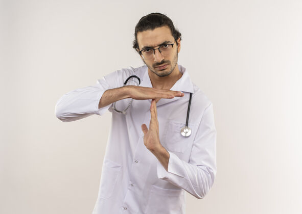 男性严格的年轻男性医生 戴着眼镜 穿着白色长袍 听诊器在隔离的白色墙壁上显示超时手势 并留有复印空间严格光学手势