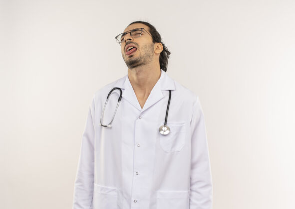 男性闭着眼睛戴着眼镜的年轻男医生穿着白色长袍 听诊器在隔离的白色墙壁上显示舌头 并留有复印空间年轻舌头穿