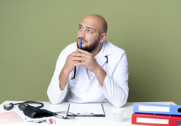 男性想着年轻的秃头男医生穿着医用长袍和听诊器坐在办公桌前拿着医疗工具把笔放在嘴上隔离在绿色的墙上医疗笔长袍
