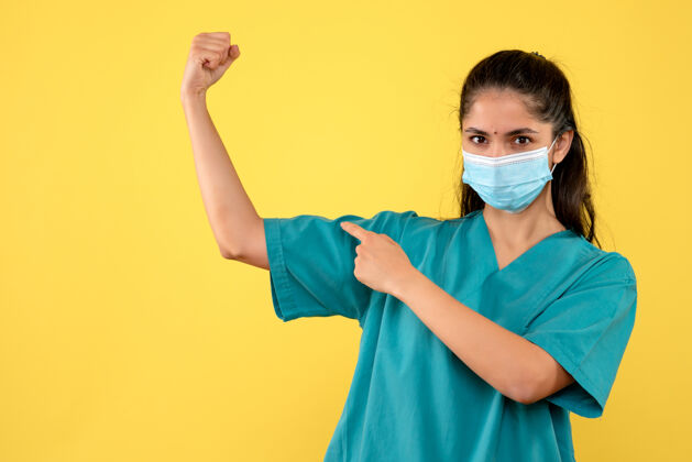 肖像黄色墙壁上 戴着医用面罩的漂亮女医生正对着她的手臂肌肉指成人护士