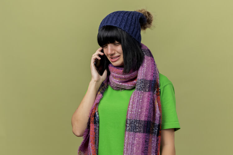 围巾戴着冬日帽子和围巾的悲伤的年轻白种人女孩在电话里说话 在橄榄绿的背景下看着孤立的复制空间帽子疾病女孩