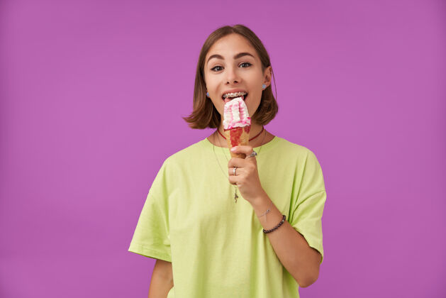 女士一个漂亮漂亮的女孩在紫色的墙上舔着冰淇淋 穿着绿色t恤 戴着牙套 手镯 戒指和项链休闲年轻手镯