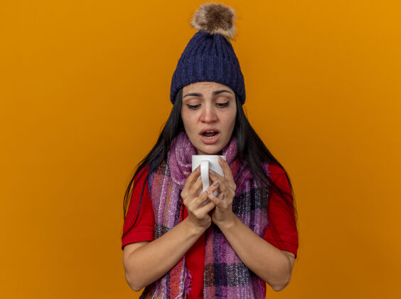 茶印象深刻的年轻白种人生病的女孩戴着冬天的帽子和围巾拿着一杯茶看里面的杯子孤立的橙色背景与复制空间帽子病抱着