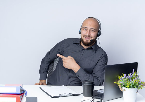 坐着令人印象深刻的年轻秃头呼叫中心男子戴着耳机坐在办公桌旁 工作工具指向隔离在白色的一边办公桌中心年轻