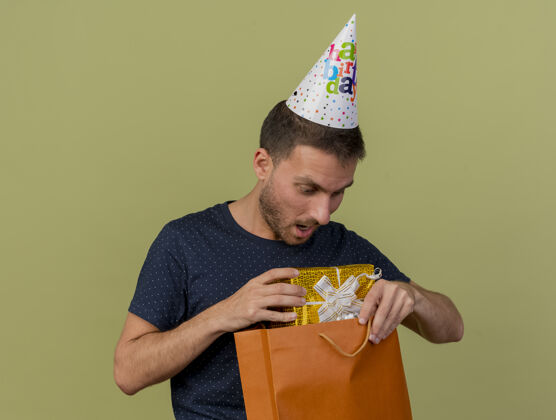 帽子一个戴着生日帽的英俊白人男子拿着礼物盒 看着一个孤立在橄榄绿背景上的纸购物袋 还有一个复制空间购物惊喜持有