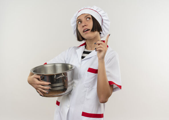 女印象深刻的年轻女厨师在厨师制服举行锅举起手指 抬头看白色与复制空间隔离锅复制向上