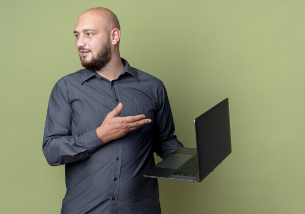 人令人印象深刻的年轻秃头呼叫中心男子举行 并用手指着笔记本电脑 看着一边孤立的橄榄绿与复制空间年轻电话印象