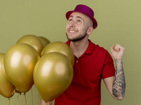 气球快乐的年轻英俊的斯拉夫党人戴着党的帽子站在气球旁抬头做是的手势孤立在橄榄绿的背景年轻帽子快乐