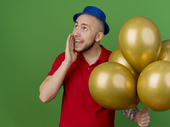 耳语令人印象深刻的年轻英俊的斯拉夫党人戴着党的帽子拿着气球看着一边低语绿色背景孤立派对气球小伙子