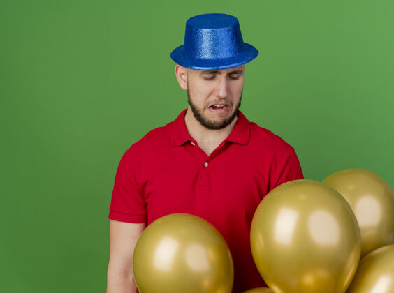 站着年轻英俊的斯拉夫党人戴着党的帽子站在气球后面看着他们在绿色背景与复制空间孤立年轻帅哥年轻人小伙子派对