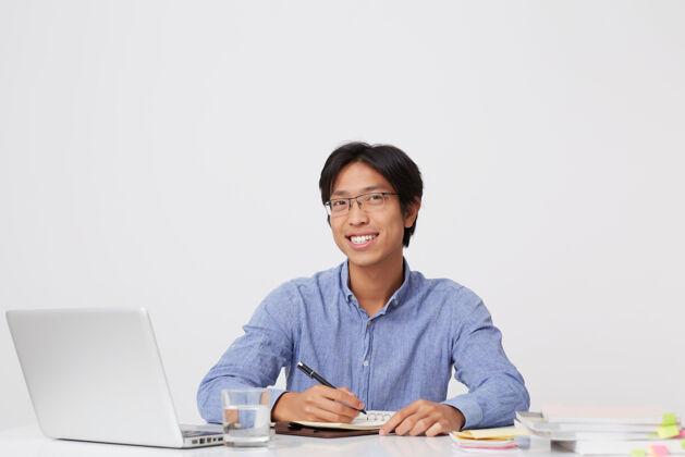 办公桌快乐英俊的亚洲年轻商人戴着眼镜在笔记本上写字在白墙上用笔记本电脑工作桌子写作英俊