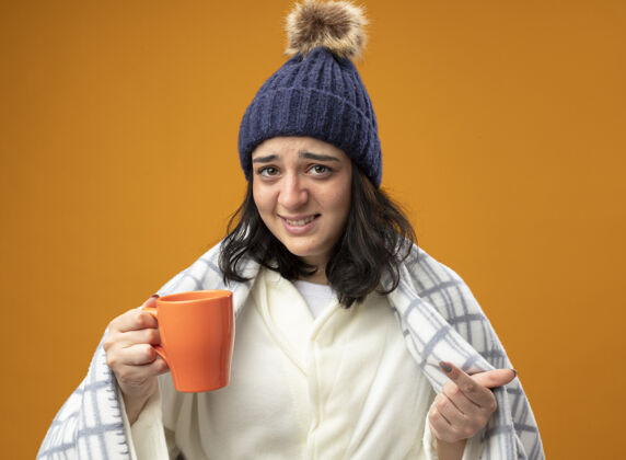 长袍恼怒的年轻白种人生病的女孩穿着长袍 戴着格子呢的冬帽 手里拿着一杯茶 看着和橙色背景隔离的摄像机帽子格子年轻