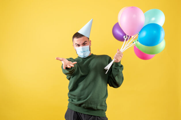快乐正面图：黄色背景上戴着面具手持彩色气球的年轻男性圣诞节生日气球