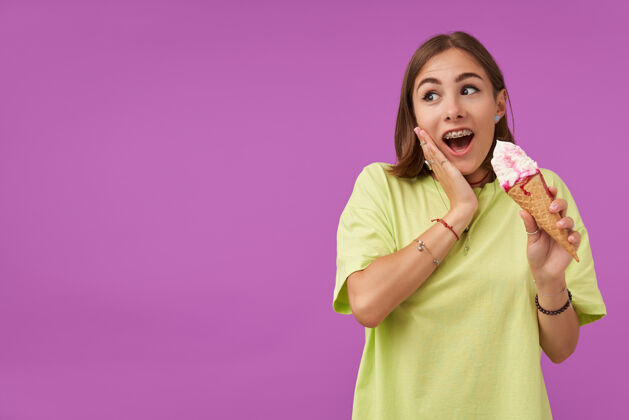 单独女学生 手里拿着冰激凌的年轻女士 摸着脸颊 震惊地看着紫色墙壁上的复制空间穿着绿色t恤 戴着戒指和项链圆锥体牙套惊喜
