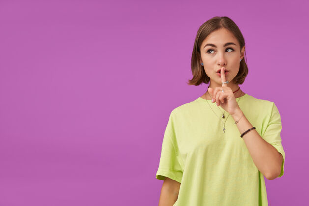 年轻漂亮漂亮的女孩的肖像用手指触摸嘴唇 显示一个沉默的标志在紫色墙上的复制空间看左边穿着绿色t恤 手镯和戒指深色手指短发