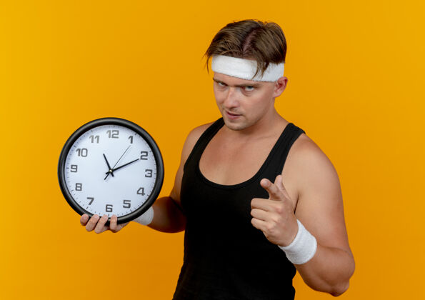 英俊自信的年轻英俊的运动型男人戴着头带和腕带拿着时钟 孤立地指着橙色橙色时钟腕带