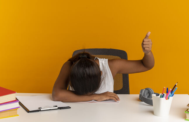 女学生年轻的女学生低着头坐在书桌旁 手里拿着学习用具 她的大拇指孤零零地竖在橙色的墙上年轻书桌拇指