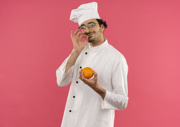 请年轻的男厨师穿着厨师制服 戴着橘黄色的眼镜 在粉红色的墙壁上展示着独特的美味姿态展示厨师美味