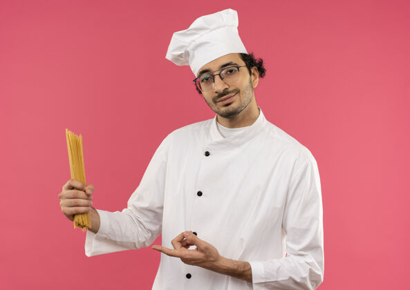 意大利面面带微笑的年轻男厨师身穿厨师制服 手持眼镜 指着隔离在粉红色墙上的意大利面点拿着厨师
