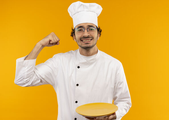 年轻面带微笑的年轻男厨师穿着厨师制服 戴着眼镜 举着盘子 做着坚强的手势孤立在黄色的墙上微笑手势强壮
