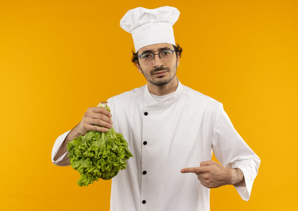 穿高兴的年轻男厨师穿着厨师制服 拿着眼镜 指着隔离在黄色墙上的沙拉点请男