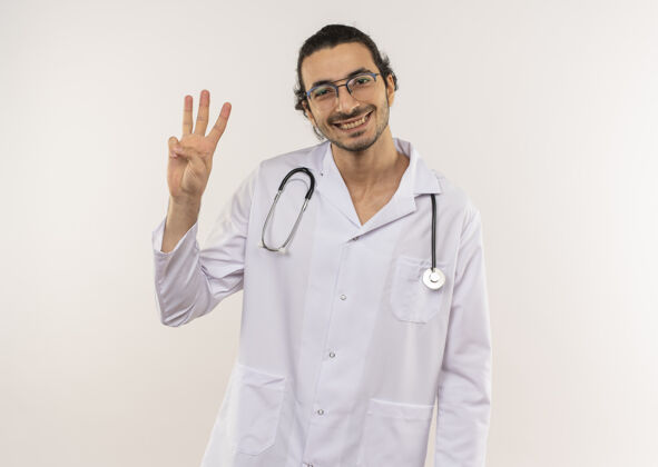 微笑微笑的年轻男性医生 戴着眼镜 穿着白色长袍 听诊器 在隔离的白墙上显示三个 还有复印空间表演穿眼镜
