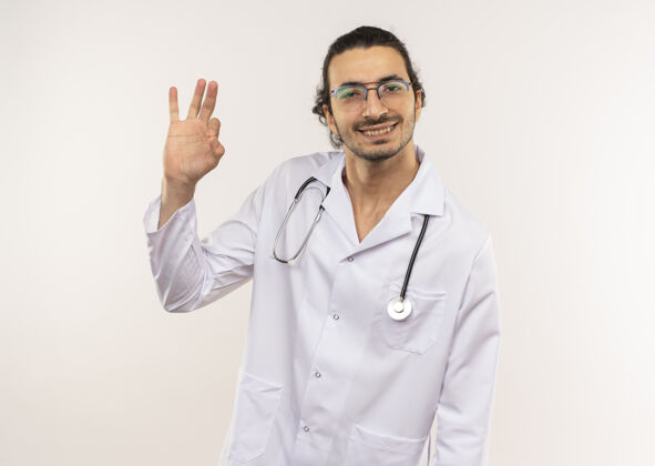 穿微笑的年轻男性医生 戴着眼镜 穿着白色长袍 听诊器 在隔离的白色墙壁上显示出良好的姿势 并留有复印空间手势展示光学
